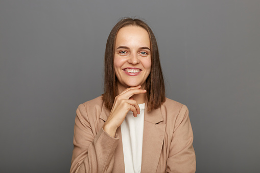 Indoor shot of smiling positive woman wearing beige jacket standing isolated over gray background, looking at camera with happy expression, keeps hand on chin, expressing positiveness.