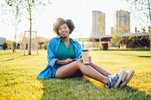 Attractive dark skinned hipster girl in casual wear sitting at green grass recreating on sunny day, smiling beautiful african american woman spending weekends in city park enjoy free time