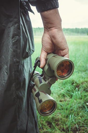 closeup view of hand of huntsman or traveller or border guardian  in dark khaki raincoat holding green binoculars with red lens against background of grassy field in forest or swamp