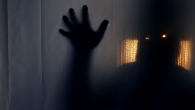 Silhouette of a man in a hood with glowing luminous eyes behind a transparent curtain, pulling his hand into the camera. The concept of otherworldly creatures, demons, paranormality, ghosts. 4k video 60 fps.