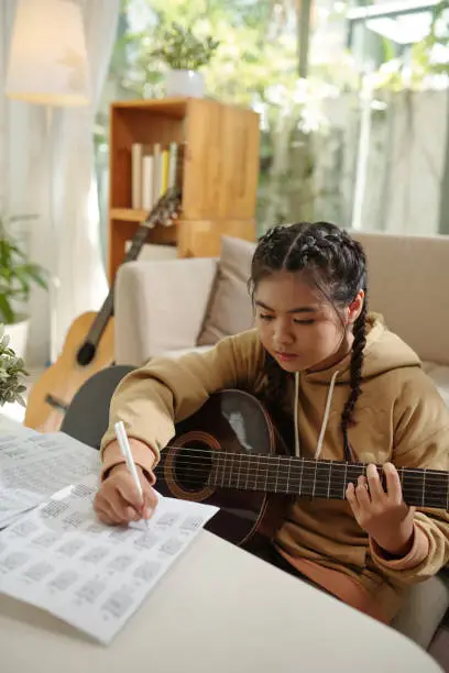 Concentrated teenage girl writing down music notes when playing guitar at home