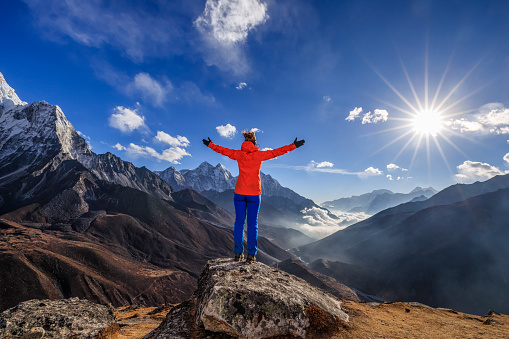 Young woman, wearing red jacket, lifts her arms in victory. She is standing on the top of a mountain and watching sunset over Himalayas .Mount Everest National Park. This is the highest national park in the world, with the entire park located above 3,000 m ( 9,700 ft).