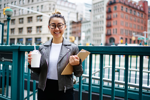 Half length portrait of cheerful female student with sketchbook and takeaway milk shake in hands standing at urbanity area and smiling at camera, happy Asian woman in tredny spectacles posing outdoors