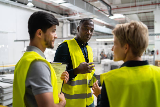 istock Three factory workers wearing reflective jackets talking together with the production line behind 1461457604