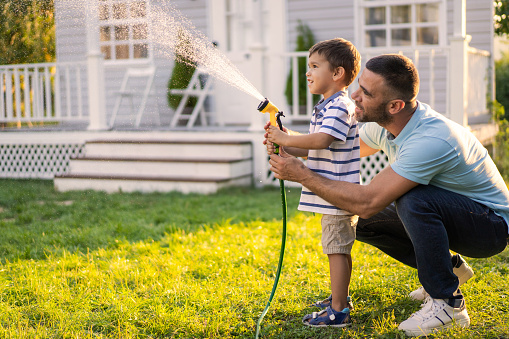 A smiling father is helping his son to hold a garden hose as they spray water all over the grass outside on a sunny day