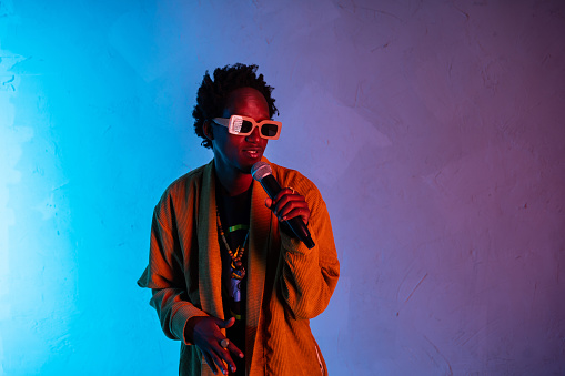 Portrait of an African-American man standing looking away from camera and holding a microphone while singing, he is wearing street style clothing and sunglasses against gradient purple to blue background