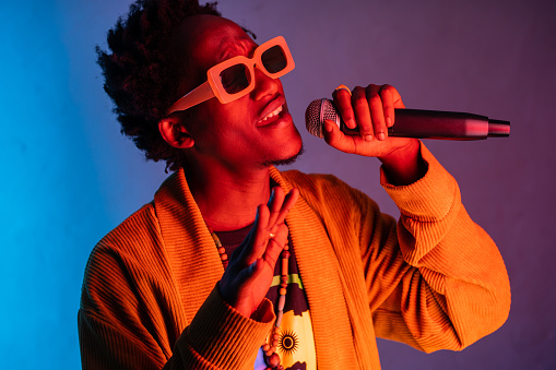Portrait of an African-American man singing into a microphone, wearing street style clothing and sunglasses on purple blue gradient background