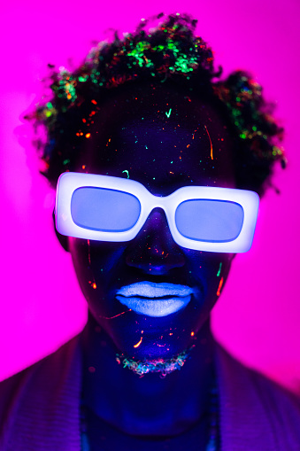 A headshot of an unrecognizable man with glasses and lips lit by ultraviolet lights and fluorescent sparkles in his hair and chin against bright pink background