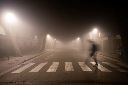 Defocused man walking on crosswalk on a foggy night in city.