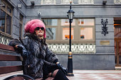 Fashionable pretty woman in sunglasses wearing natural fur coat and pink fur hat sitting on bench in city district, looking away. Stylish lady posing outdoor. Fashion style concept. Copy space for ad