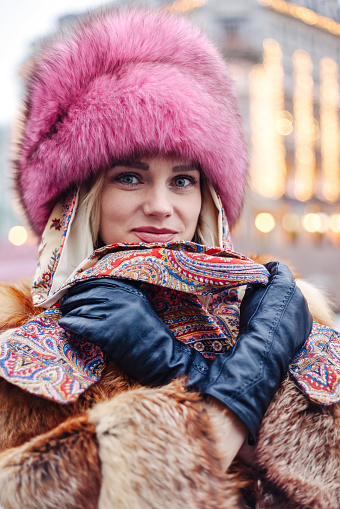 Stylish smiling woman in natural fur coat and pink fur hat posing at winter urban street, looking at camera. Portrait of fashionable lady outdoors. Fashion style concept. Copy empty text space for ad