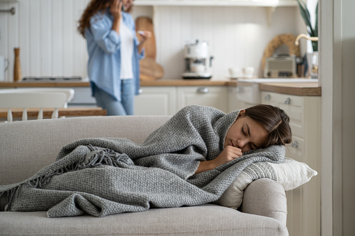 Sick repressed teen girl sleeping in house on couch wrapped in blanket warming up in cold weather. Caring woman calling by phone holding thermometer standing behind schoolgirl lying with closed eyes