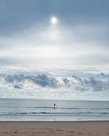 A man standing on stand up paddleboard by the sea in the morning on a cloudy day with the sun coming out.
