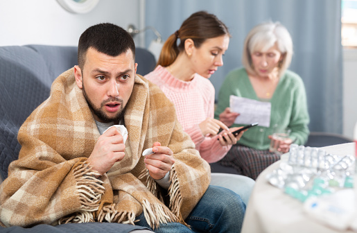 Sick man sneezing in napkin, young and elderly anxious women with medicines sitting in livingroom