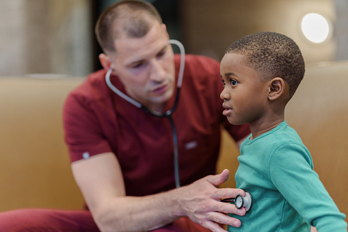 A male nurse uses a stethoscope to check the heart and lungs of an elementary age African American boy during an annual check-up in a medical clinic or hospital.