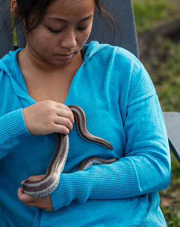 A young Asian woman in her 20s sitting outdoors in the back yard, holding her pet snake. It is a Rosy Boa, a small docile constrictor native to southwestern North America.