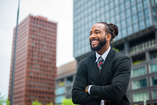 Young male African American economist leads team of professional executives to develop strategies against the crisis. He looks confident with his arms crossed. He is smiling and looking away. Defocused city buildings in the background.