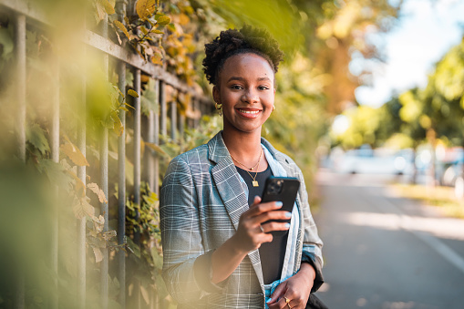 Young African American woman commuting in the city, using a smart phone and texting. She is wearing casual clothes on a sunny day.