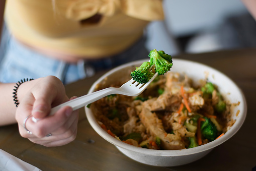 Young woman eating take out food with closeup on a broccoli on a plastic disposable sigle use fork