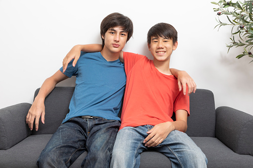 Two happy Asian and caucasian teen boy sit on a sofa at home. Two friend happy and sit on sofa.