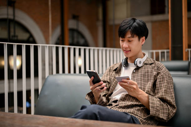 Happy Asian man holding a smartphone and a credit card, using mobile banking application Happy Asian man in casual clothes holding a smartphone and a credit card, using mobile banking application, registering credit card payment on shopping app. asian man headphones stock pictures, royalty-free photos & images
