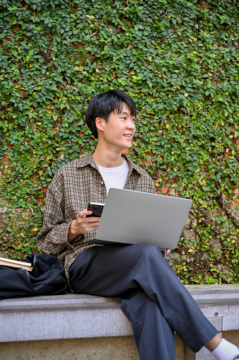 Portrait of a young Asian man in trendy outfit remote working in the city garden park, using laptop computer, sipping coffee, sitting on a bench.