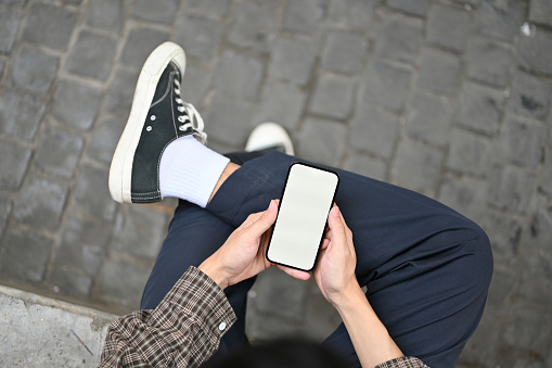 Top view of a man using his smartphone, chatting with someone or using mobile application while relax sitting at the city square street. phone white screen mockup