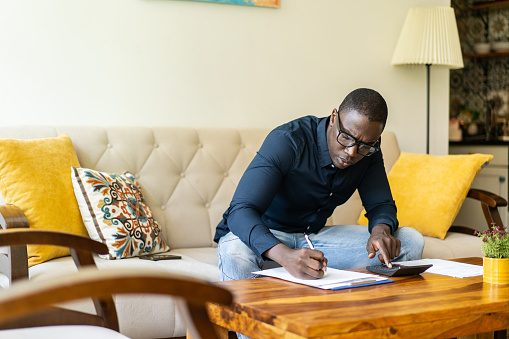 An African-American man is calculating his home finances leaning forward to work on a coffee table at home in his living room
