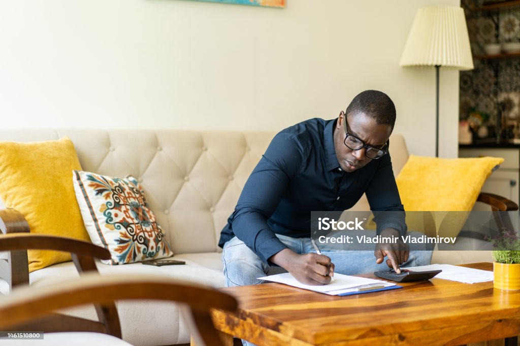 A worried man is adding up his household expenses An African-American man is calculating his home finances leaning forward to work on a coffee table at home in his living room Price Stock Photo