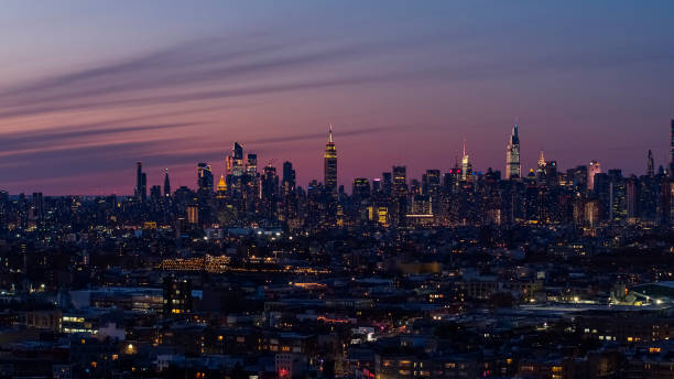 Remote view of the Manhattan Skyline illuminated in the night over the residential district of Bushwick, Brooklyn, in the sunset. manhattan midtown skyline incluye el empire state building, hudson yards y otros rascacielos icónicos. vista sobre el distrito residencial de bushwick, brooklyn, al atardecer, con el espectacular cielo colorido. panorama cosido extra grande y de alta reso