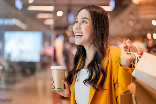 surprising cheerful asian female adult woman shopper hand hold shopping bag and coffee paper cup walking buying cloths in department store shopping mall with happiness exited toothy smiling joyful