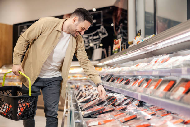 A cheerful man is choosing meat in supermarket while holding shopping cart. stock photo