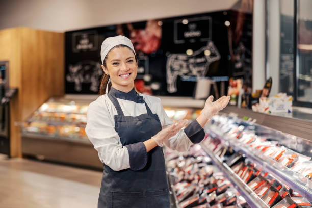 um açougueiro feliz está aparecendo ao redor do departamento de carne enquanto está no supermercado e sorri para a câmera. - supermarket meat store manager - fotografias e filmes do acervo