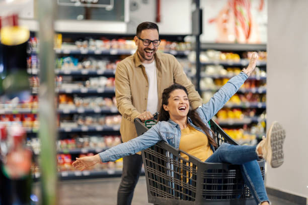A happy man drives his woman in shopping cart in supermarket. stock photo