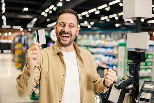 A smiling man holding credit card and celebrating while smiling at the camera in supermarket.