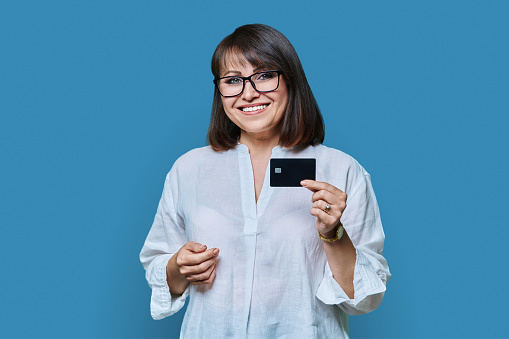 Attractive middle aged smiling woman holding plastic credit card, on blue color studio background. Confident positive mature business female worker manager customer bank staff looking at camera