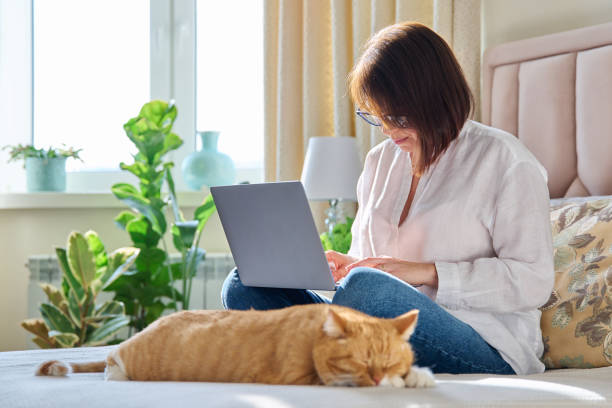 istock Mature woman using laptop sitting on couch at home, with relaxed sleeping ginger cat 1461543531