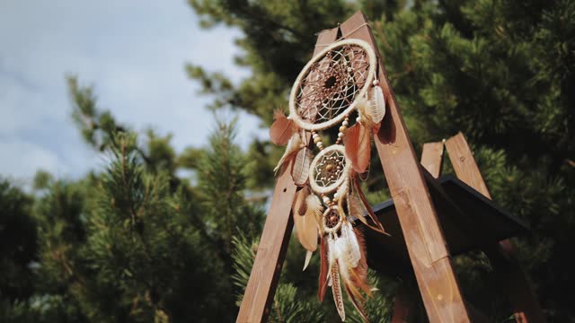 Wedding decoration with wooden dreamcatcher with brown feathers close-up shot. Feathers swaying in the wind, slow motion shot. Pine trees background.
