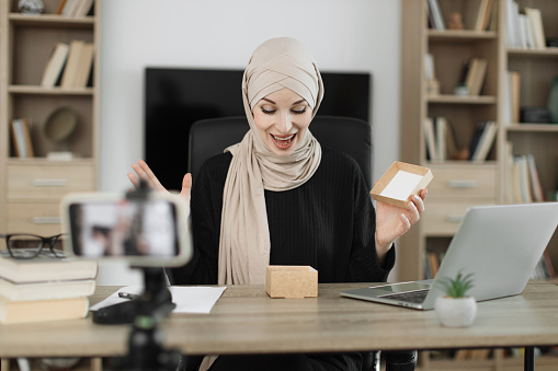 Attractive muslim female blogger unpacking gift boxes presents from companies and doing live streaming. Arab woman using smart phone camera for creating content.