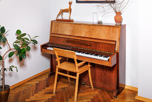 An old orange-coloured piano with a lacquered surface sits next to a ficus flowerpot in the corner of an empty room with white walls.