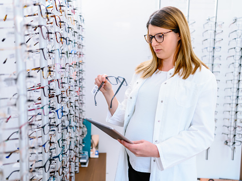 Optometry seller woman showing eyeglasses assortment on the display shelf showcase in the optical shop, picking up frames, a doctor helping the client to make choice with spectacles