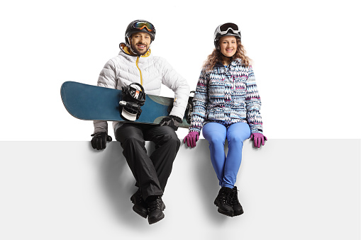 Young man and woman wearing winter equipment sitting on a blank panel and holding a snowboard isolated on white background