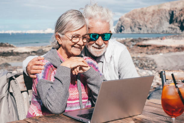 lächelndes älteres paar, das zusammen auf dem laptop surft und den sonnigen tag im freien am strand genießt, sitzt an einem holztisch mit orangefarbenem cocktail. ältere menschen im urlaub oder ruhestand auf see - bonding horizontal surfing surf stock-fotos und bilder