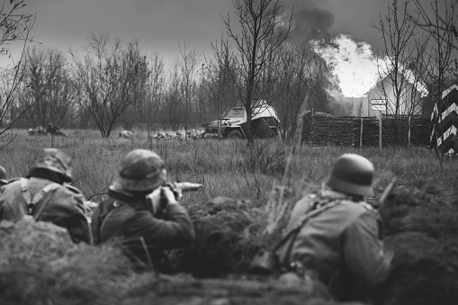 Re-enactors Armed Rifles And Dressed As World War Ii German Wehrmacht Infantry Soldiers Fighting Defensively In Trench. Fight Against Combat Vehicle. Building On Fire On Background. Black White Colors