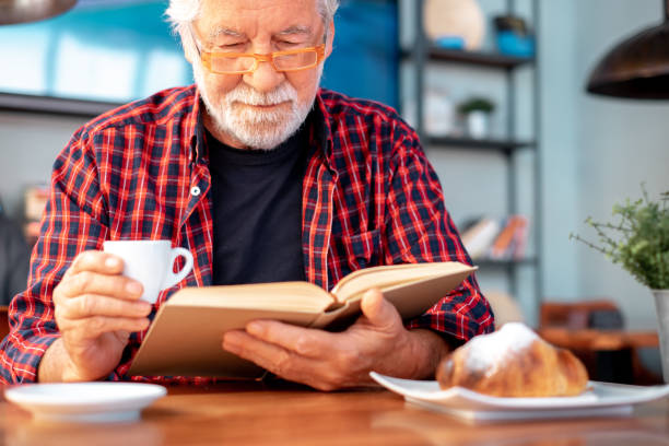 istock Smiling senior bearded man sitting at cafe table with a book while enjoying reading pleasure and breakfast with espresso coffee cup and croissant. Elderly caucasian male in eyeglasses and checkered shirt 1461576896