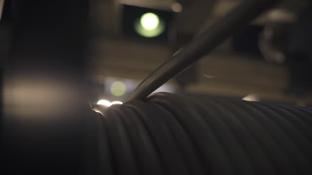 The cable slowly unwinds from a large spool at the cable factory. Close-up. A bright lamp in the background shines brightly. Production process at the cable factory. 4K
