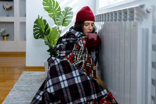 A young woman is feeling cold at home because of an energetic crisis and trying to warm herself up while sitting near the radiator and drinking a hot tea.