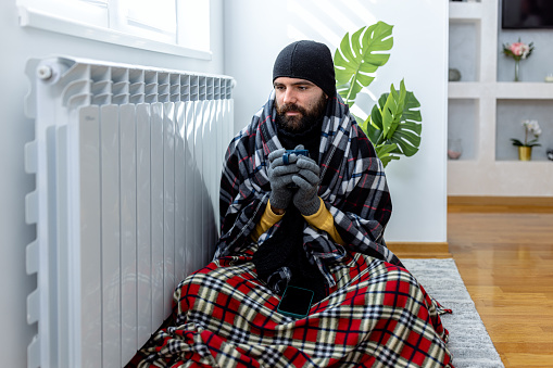 A man in warm clothing is shivering and wearing a blanket and freezing at home while drinking a hot cup o tea and sitting near a radiator.