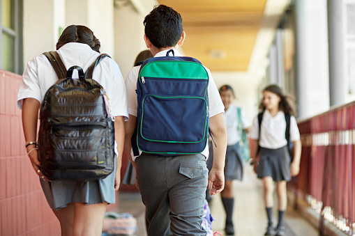 Focus on foreground elementary-age boy and girl carrying backpacks and moving along outdoor hallway side by side.