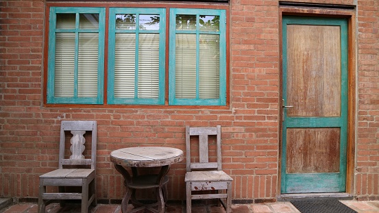 Front view of Indonesian old Vintage exterior house. Wooden chair, round table and wooden door. Old design. Green windows on the brown brick wall. square brick pattern on the floor.